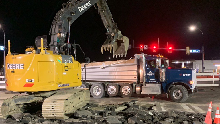 A nighttime photo shows an excavator moving broken pieces of a street into a dump truck.