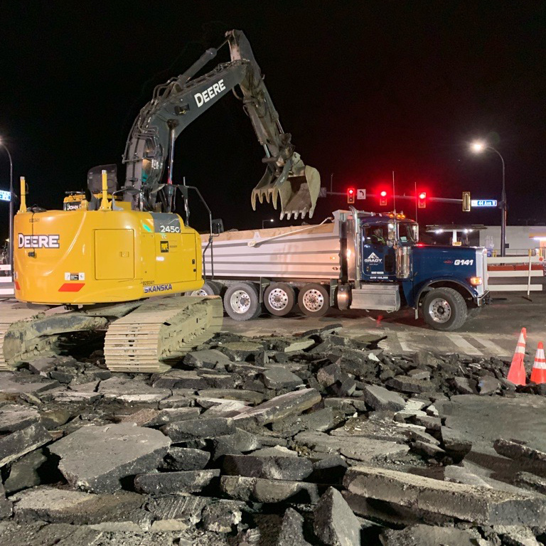 A nighttime photo shows an excavator moving broken pieces of a street into a dump truck.