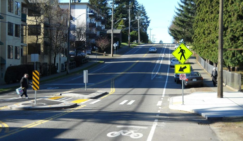 An image of a roadway with a bike lane, as well as a raised pedestrian crossing island. The crosswalk has white painted stripes and is elevated, leading to a pedestrian refuge island in the center of the road, marked with yellow and black striped barriers. On the right side, there is a yellow pedestrian crossing sign.