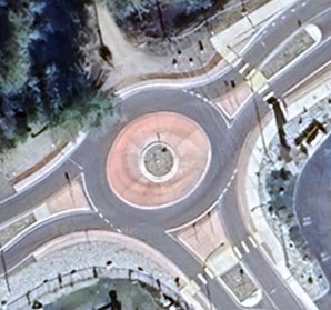 An overhead view of a three-legged roundabout that has one lane for each direction. The roundabout has a landscaped island, and sidewalk crossings on all two of the three legs, one is not shown in the image. There are mountable truck aprons in the center of the roundabout, adjacent to the landscaped island.