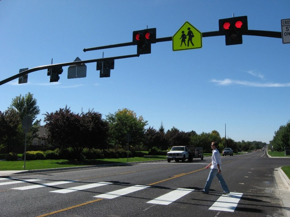 An image showing an example of a three-lane road, one in each direction and a two-way center left-turn lane. There is an overhead pedestrian hybrid beacon with solid red lights activated. A pedestrian is crossing the roadway, using the crosswalk and a vehicle is stopped at a stop line.