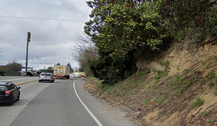 An image showing two lanes of westbound SR 900, west of 133rd Street South, next to a steep hill. There is grass vegetation, trees, and exposed dirt on the steep hill. There is a narrow shoulder between the travel lanes and the steep hill with small accumulations of dirt on the shoulder.