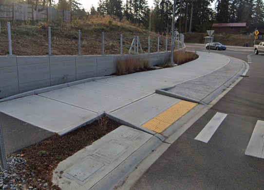 An image showing an example of a wide shared-use-path that is protected with a small (4 to 6 feet high) retaining wall that is built into a hill. A patterned concrete buffer separates the shared-use-path and travel lanes until the crosswalk. On the near side of the image, a vegetated buffer separates the shared-use-path and travel lanes.