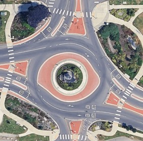 An overhead view of a four-legged roundabout that has two lanes in the east and west direction, and one lane in the north and south directions. The roundabout has a landscaped island and sidewalk crossings on all four legs. There are mountable truck aprons in the center of the roundabout, adjacent to the landscaped island.